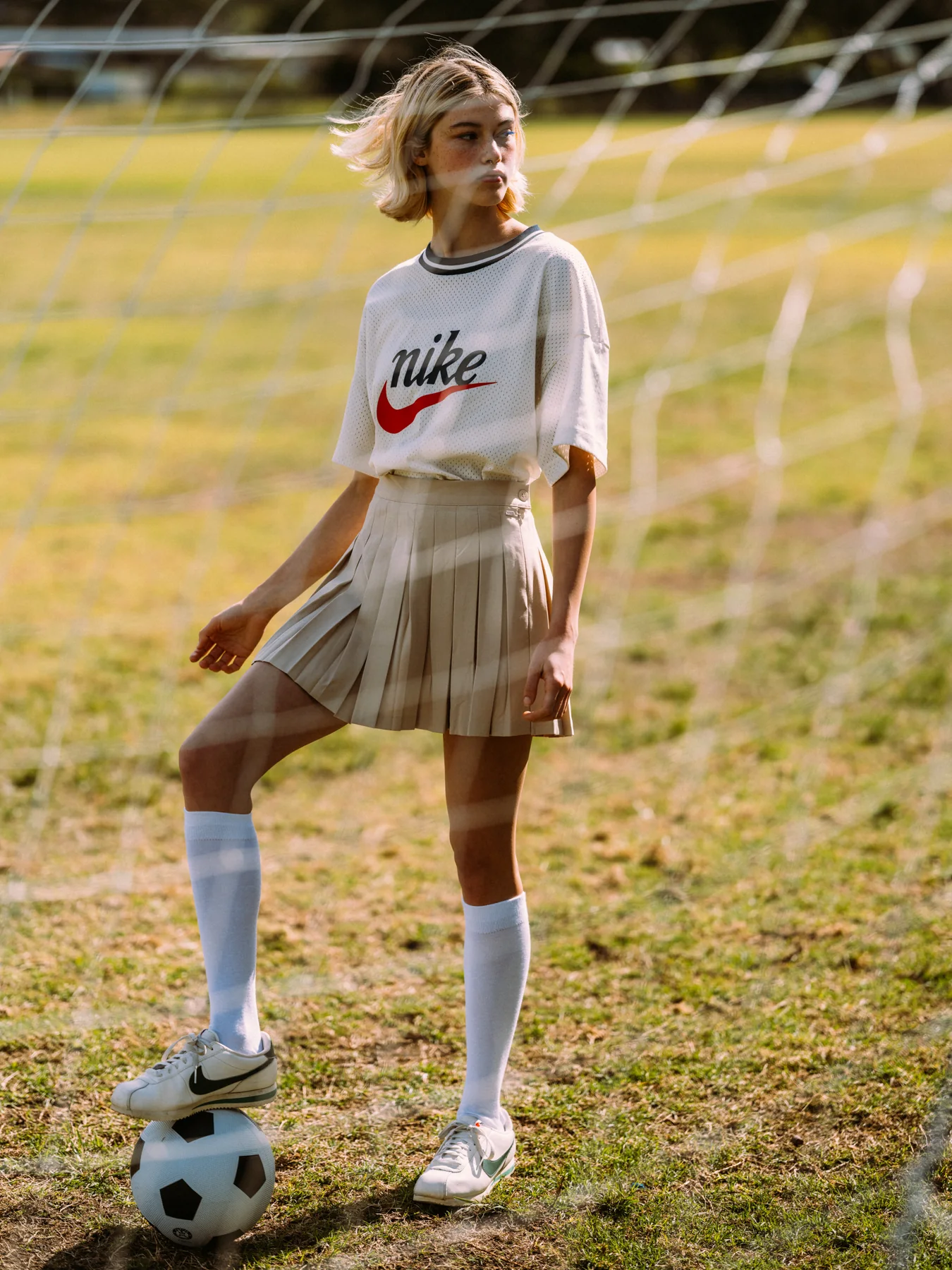 Gracie Rae in a white T-shirt and pleated skirt with one foot on a football beside a goal