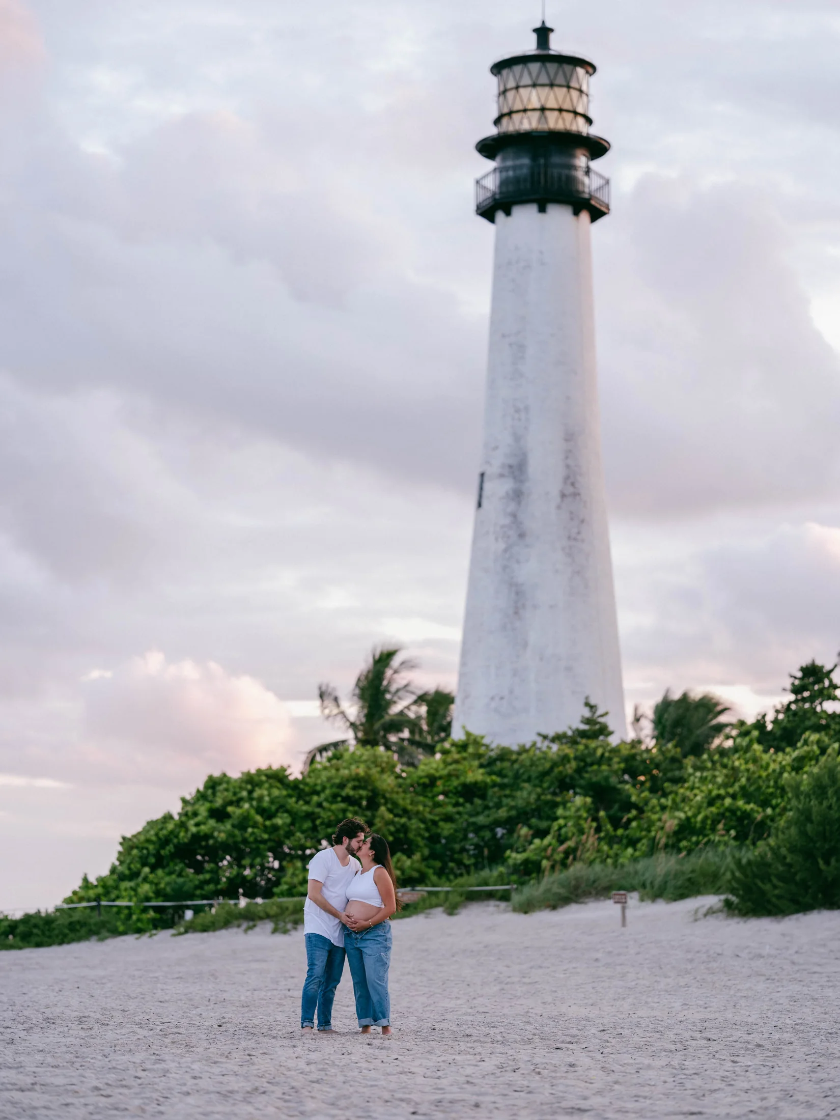 Peter and Michelle embracing on the beach below a white lighthouse