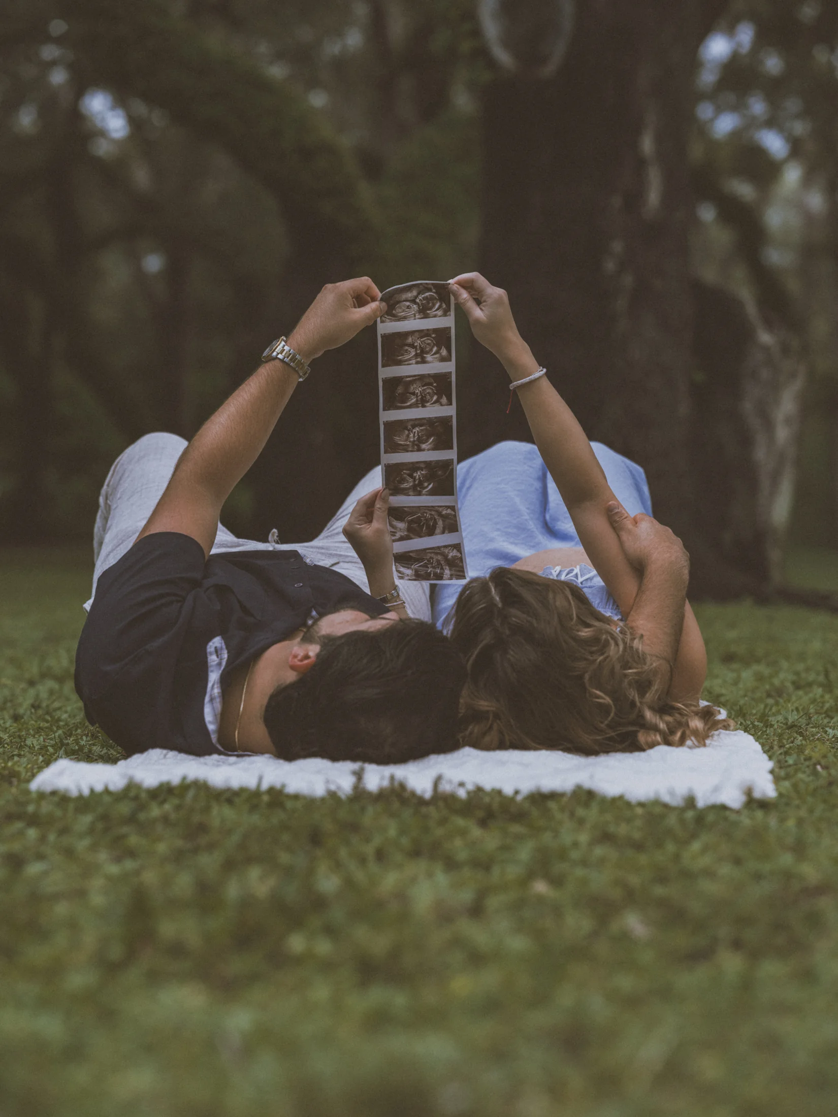 Kelsey and her partner lying on a blanket in the park holding up ultrasound photographs