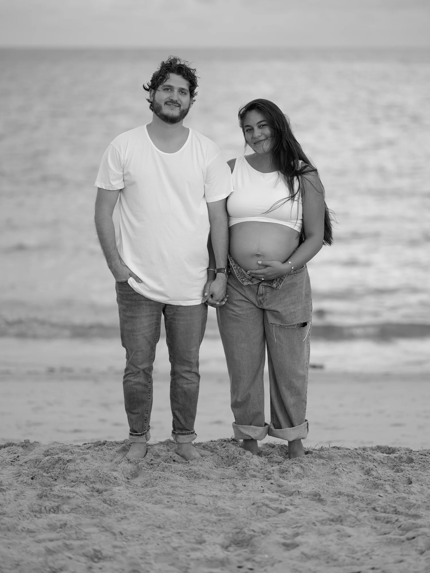 Black-and-white maternity portrait of Peter and Michelle standing together beside the sea