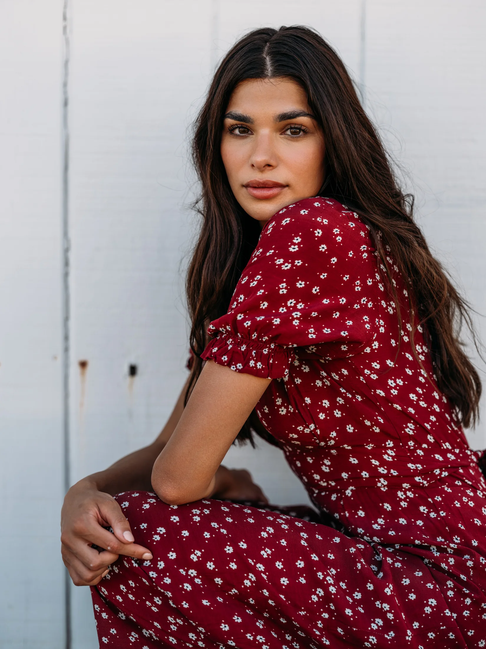 Rhea Rai looking over her shoulder in a red floral dress against a white wall