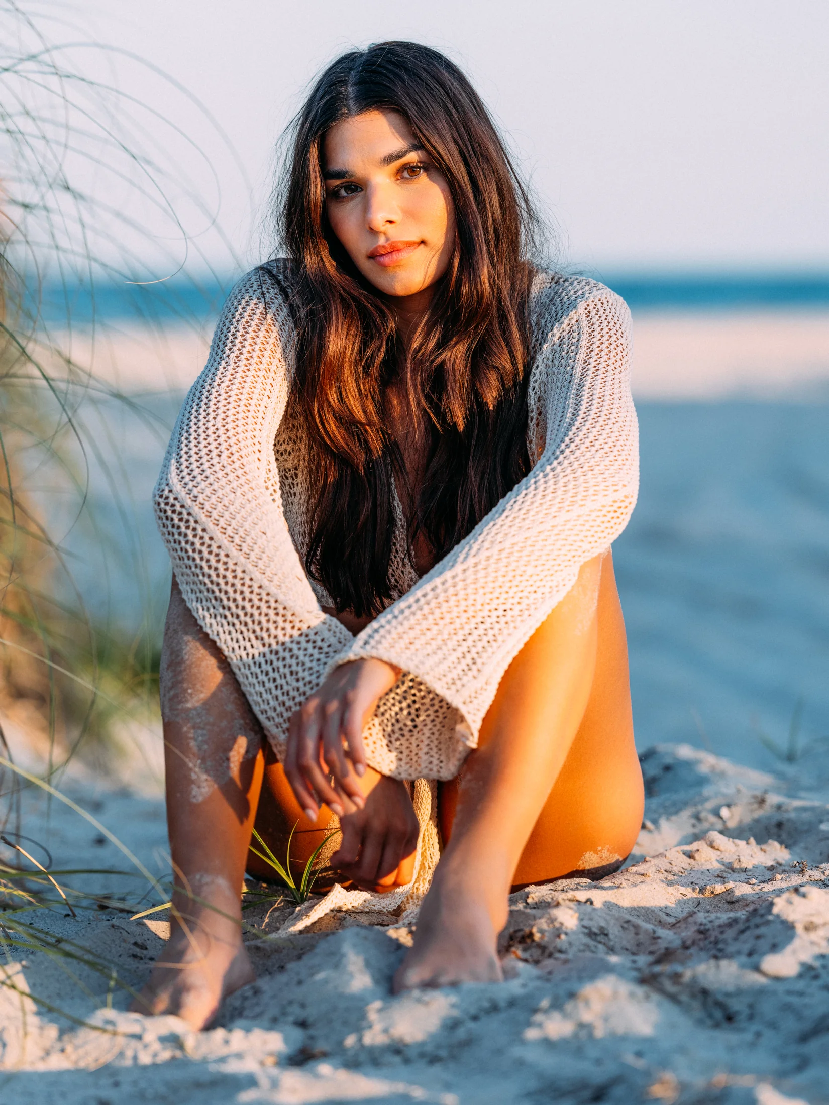 Rhea Rai seated on a sandy beach in a loose white knit top
